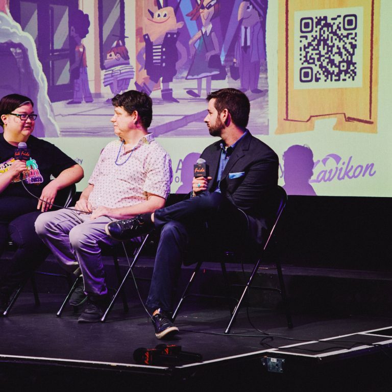 Alex Astrella [Right] and two other panelists sit on folding chairs on a stage during a Q&A, two holding microphones, in front of a projection screen showing cartoon characters and sponsor logos.