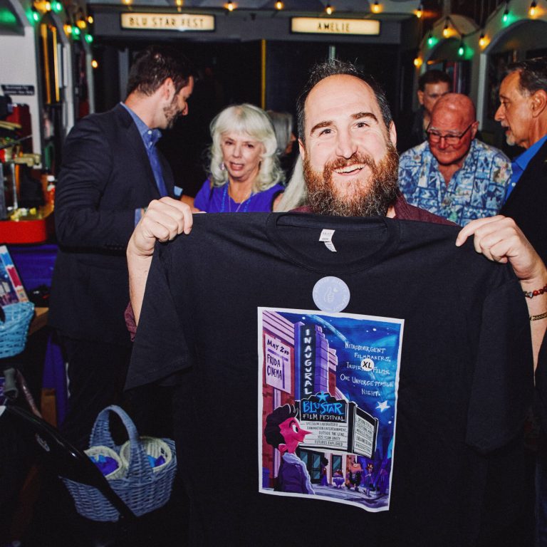 A bearded man smiles as he holds up a black XL T-shirt printed with the colorful "Bluestar Film Festival" poster, surrounded by a group of people at the event.