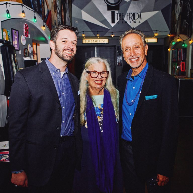 Three people pose for a photo at an indoor event: Alex Astrella, a bearded man in a navy blazer and blue shirt on the left, his Mother with long blonde hair, red-framed glasses, and a purple scarf in the center, and his Father in a black blazer and blue shirt on the right. Behind them, festive string lights hang across a hallway decorated with posters and a sign reading "The Frida Cinema," with a smaller sign above a doorway that says "BLU STAR FEST."