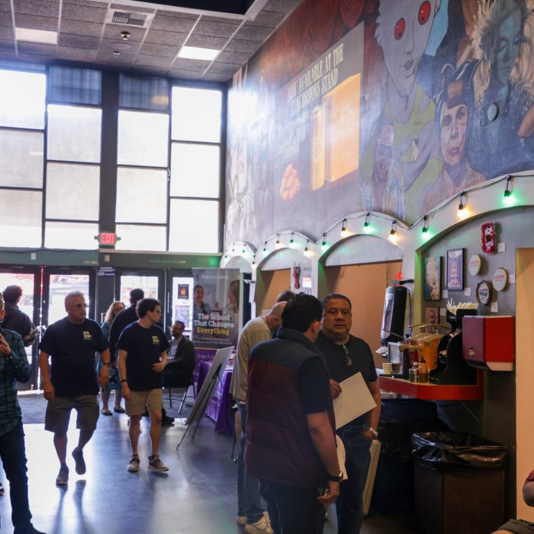 Inside the Frida Cinema lobby during the Bluestar Film Festival, numerous people walk and converse in a wide, open space. Natural light floods in through tall rectangular windows and glass entrance doors marked with an illuminated red EXIT sign. In the foreground on the right, a man in a red polo shirt sits looking at a film reel, while behind him a blue banner reads "EXPANDING EMPL[OYMENT] OF AUTISTIC TALE[NT IN THE] ENTERTAINMENT I[NDUSTRY]." Further back, a concession stand with a red counter sits beneath a massive mural depicting movie characters, including the alien from Fantastic Planet. Festive string lights hang along the arched alcoves, and attendees in casual clothing mill about, some holding papers and programs, creating a bustling, welcoming festival atmosphere.
