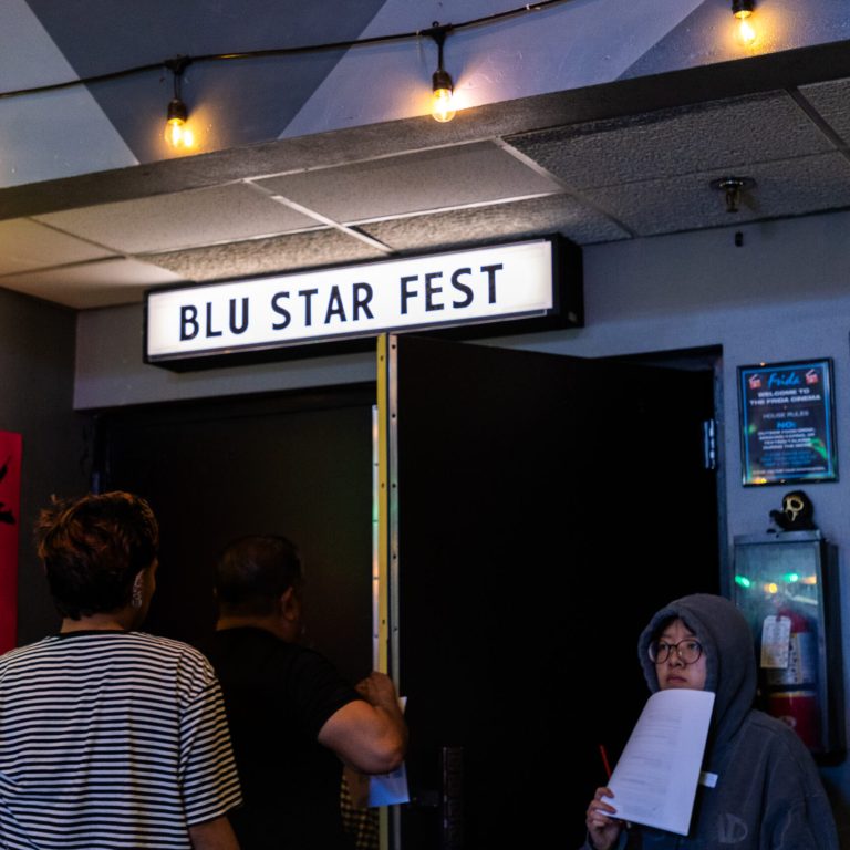 People stand in a dimly lit theater corridor beneath an illuminated "BLU STAR FEST" sign, one woman in a hoodie holding a red pen and a paper.