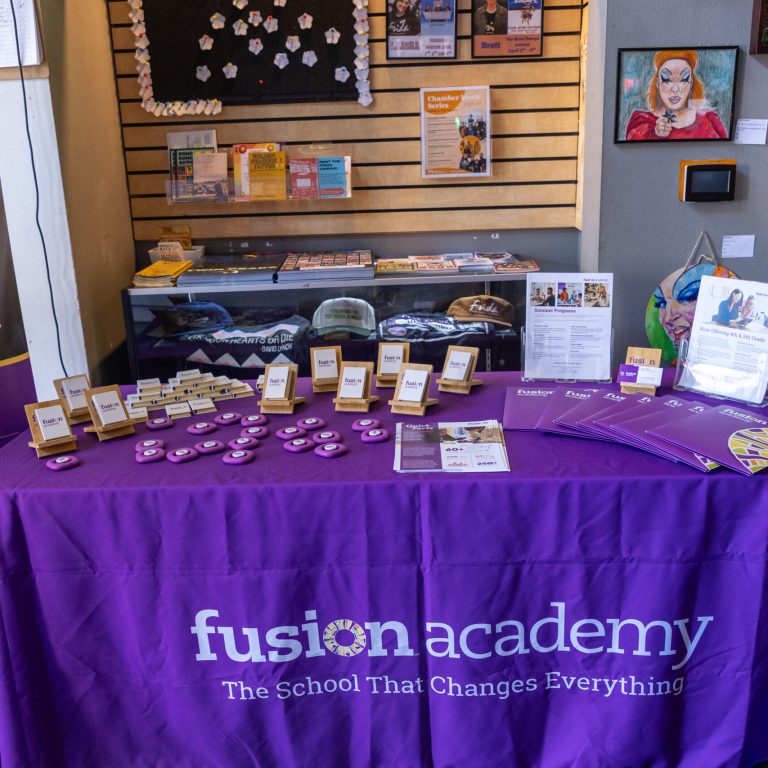 A Fusion Academy promotional table covered in a purple branded cloth displays brochures, buttons, and small signs at the Blu Star Film Festival.
