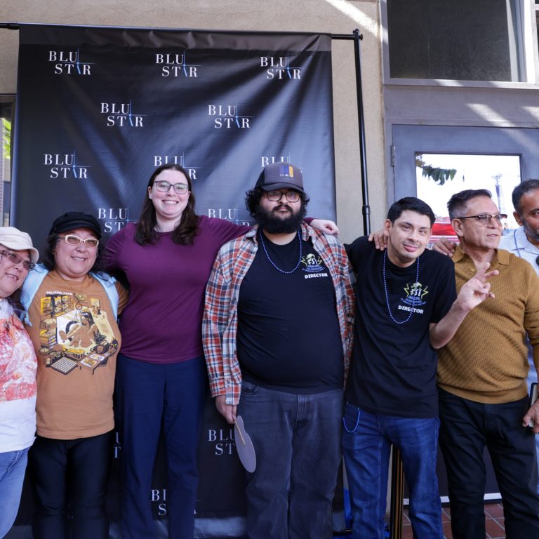 Six people stand together for a group photo on the sidewalk outside the Frida Cinema, posing in front of a large black step-and-repeat banner printed with the repeating "BLU STAR" logo in white. From left to right: a woman in a white bucket hat and white graphic T-shirt makes a peace sign; a woman in a brown cap and tan T-shirt with a film-themed graphic smiles broadly; a tall woman in a purple long-sleeve shirt and glasses has her arm around the group; a man with a beard, glasses, and a plaid shirt over a black T-shirt holds a sign; a younger man in a black T-shirt with a "DIRECTOR" graphic and a beaded necklace flashes a peace sign; and two older men, one in a mustard sweater, the other in a light blue button-down shirt holding a program or card.