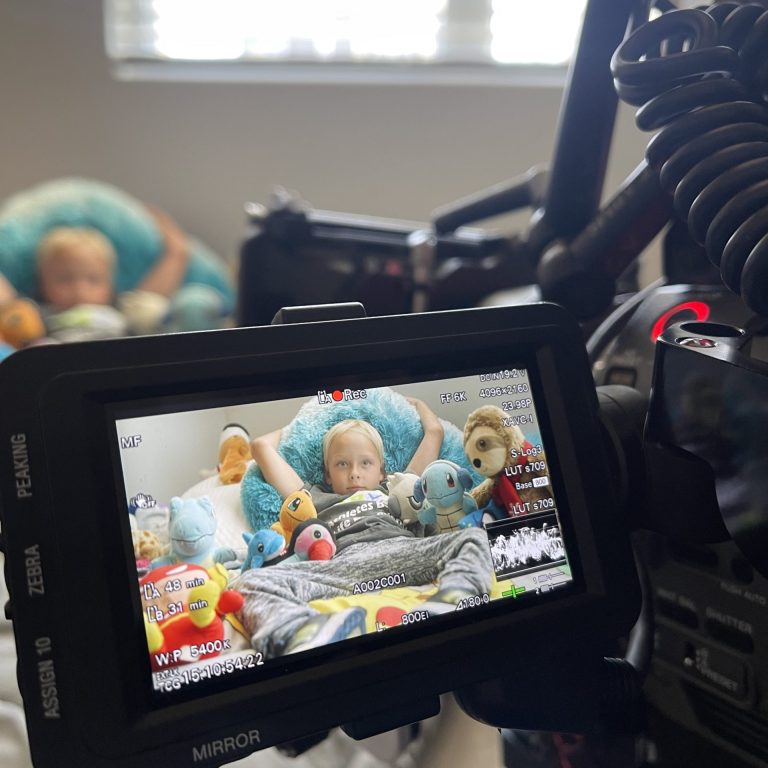Close-up of a camera monitor displaying a child lying among Pokémon stuffed animals (Snorlax, Psyduck, Squirtle, Bulbasaur, etc.), with recording overlays like "REC" and waveform.