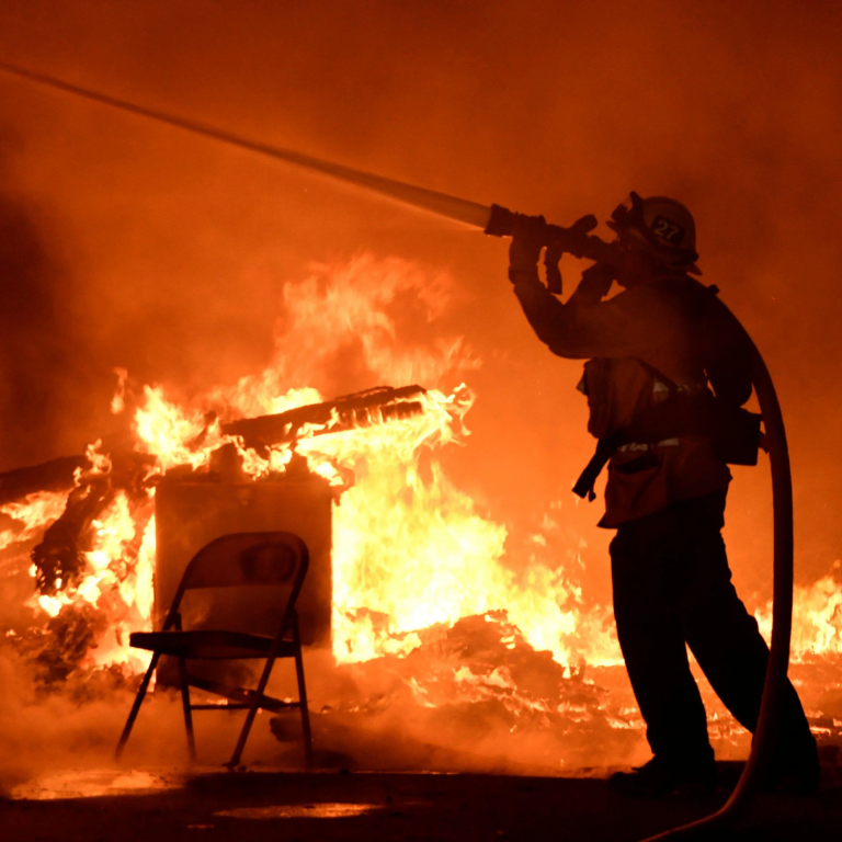 A firefighter wearing full protective gear and a helmet marked "27" is silhouetted against intense flames while directing a powerful stream of water from a hose onto a burning structure. The scene is dominated by bright orange and red hues, with thick smoke rising into the night sky. In the foreground, an empty folding chair stands near the blaze.